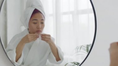 Bornoz ve havlu giymiş genç, güzel bir Asyalı kadın banyoda aynanın önünde yüzüne losyon sürerken pamuk pedleri kullanıyor.
