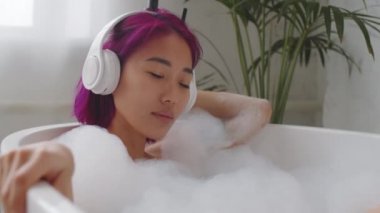 Kablosuz kulaklıklı genç, rahat Asyalı kadın küvette uzanıyor ve keyifle müzik dinliyor.