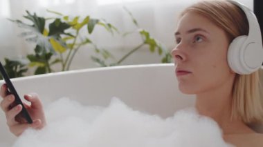 Kablosuz kulaklıklı genç kadın küvette uzanıyor, akıllı telefon kullanıyor ve gözleri kapalı müzik dinliyor.