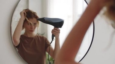 Genç ve güzel bir kadın aynanın önünde duruyor ve banyo yaptıktan sonra saç kurutma makinesi kullanıyor.