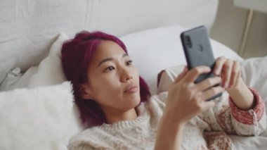 Yüksek açılı, pembe saçlı güzel Asyalı bir kadının akıllı telefonuyla mesajlaşırken ve gülümserken görüntüsü.