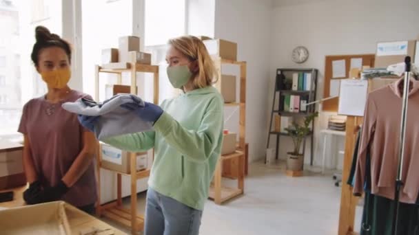 保護マスクと手袋の包装服を箱に入れ ラップトップを使用している2人の若い女性の同僚は コロナウイルスパンデミックの間にオンラインストアで出荷するための注文を準備しながら — ストック動画