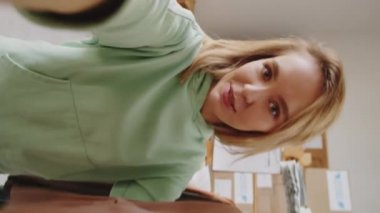 Giysi dükkanında iş saatinde müşteriyle konuşurken akıllı telefondan polo yakalı kazak gösteren ve ondan bahseden genç beyaz kadının dikey görüntüsü.