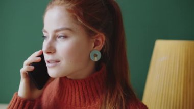 Genç kızıl saçlı bir kadının cep telefonuyla sohbet edişinin ve koyu yeşil bir duvarın önünde sırıtışının göğüs yukarı çekimi.