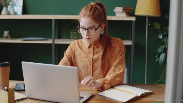 在现代办公室的办公桌前工作时 年轻的红头发女商人在笔记本电脑上打字和记笔记 — 图库视频影像