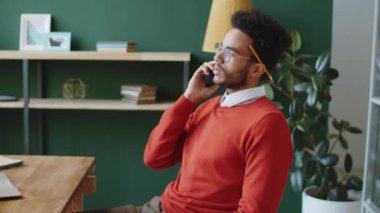 Genç melez erkek yönetici cep telefonuyla konuşuyor ve modern ofiste çalışırken notlar alıyor.