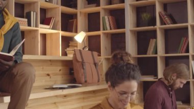 Tilting medium shot of three students sitting on benches in coworking  Afro-American guy writing in notebook, Caucasian girl typing on laptop and massaging her neck, and young man using smartphone