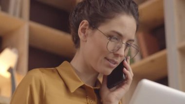 Close-up tilting down shot of lovely young Caucasian woman with curly hair, in glasses, working on laptop in cozy hipster cafe, holding smartphone to shoulder, smiling and chatting