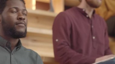 Tilting medium shot of multiethnic young students sitting on benches in warmly lit book cafe, red-haired Caucasian guy in glasses raising hand and asking question, and young girl writing in notebook