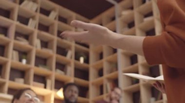 Tilting shot of female speaker giving lecture at hipster coworking cafe, and multiethnic young audience sitting on benches, looking at her, listening attentively and asking questions for discussion