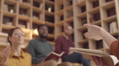 Multiethnic creative project team sitting on benches in coworking cafe, hands of speaker with notebook encouraging young Caucasian woman to speak, and male colleagues listening and making notes