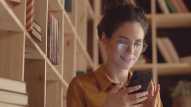 Close-up shot of lovely young Caucasian woman in glasses, with curly hair in updo, sitting in book cafe next to wooden shelves, typing message on smartphone, then looking at camera with warm smile
