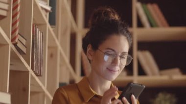 Close-up shot of smiling young Caucasian woman in glasses, with curly hair in updo, sitting in book cafe next to wooden shelves, holding smartphone, enlarging picture and typing message