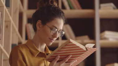 Chest-up shot of lovely young Caucasian woman in glasses, with curly hair in updo, reading book in cozy hipster cafe and turning over page, and wooden shelves in background