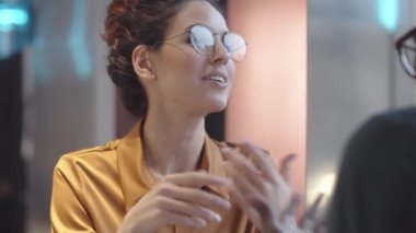 Over-shoulder chest-up shot of lovely young Caucasian woman with curly hair, in glasses sitting at table in cafe with Afro-American man, smiling, laughing and having lively conversation together