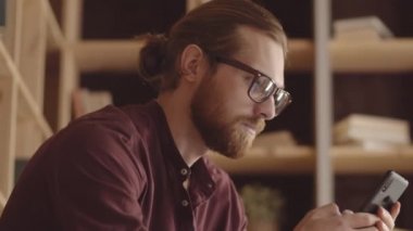 Tilting chest-up shot of red-haired Caucasian male hipster with full beard sitting in book cafe with wooden shelves, using applications on smartphone and smiling
