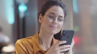 Kafede oturan, elinde akıllı telefon olan ve haberci başvurusunda biriyle sohbet eden, etrafa bakıp neşeyle gülümseyen güzel, beyaz bir kadının yakın çekimi.