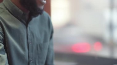 Gözlüklü, kablosuz kulaklıklı, restoranda oturan, dizüstü bilgisayarında video görüşmesi yapan, ekranda el sallayan ve konuşan Afro-Amerikan bir adamın yakın çekim görüntüsü.