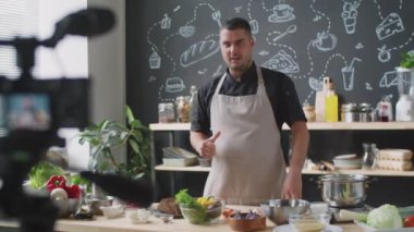 Önlüklü erkek aşçı malzemelerini gösteriyor ve mutfakta karatahta duvarla yemek vlog 'u kaydederken kameraya konuşuyor.
