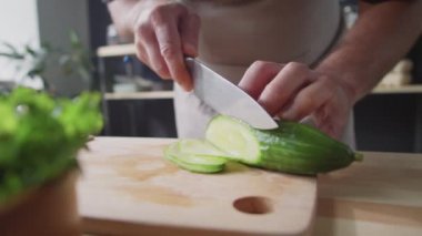 Erkek şefin, mutfaktaki tahta tahtada bıçakla taze salatalıkları ince dilimlere ayırdığı yakın çekim.