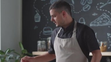 Önlüklü erkek aşçının kameraya konuşan, tavayı ocağa koyan ve mutfakta online yemek kursu verirken üzerine iki dilim ekmek atan el kamerası görüntüsü.