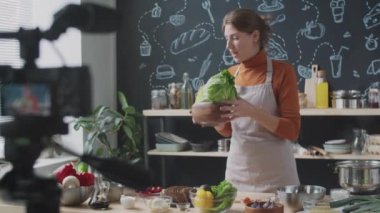 Önlüklü genç bayan yemek blogcusu taze sebzeleri gösteriyor ve mutfakta çevrimiçi yemek kursunu kaydederken kamerada konuşuyor.