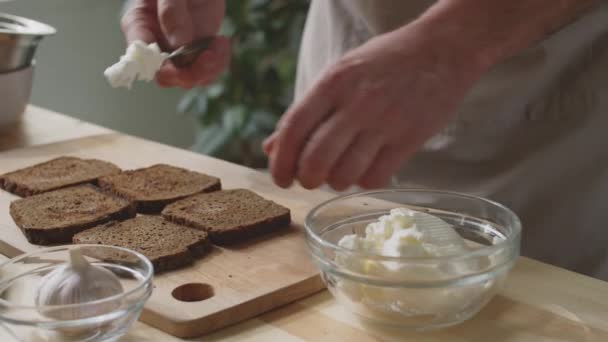 坐在围裙里的无法辨认的男性厨师将奶油奶酪放在厨房桌上烤黑麦面包片的特写镜头 — 图库视频影像