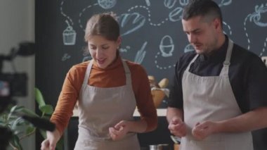 Neşeli adam ve kadın önlükleriyle mutfakta dikiliyor, kamerada yemek malzemelerini el sallıyor ve anlatıyorlar.