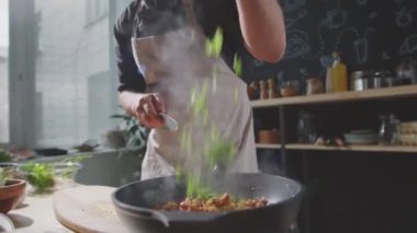 Erkek şefin apronlu orta kesiminde ete maydanoz ekleyip mutfakta yemek pişirirken sıcak tavaya yemek atarken.