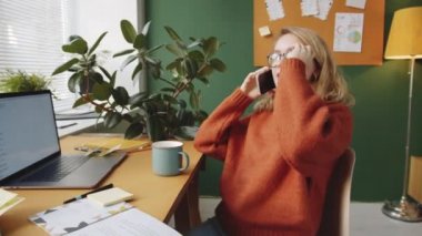 Kafkasyalı genç iş kadını ofis masasında oturuyor, cep telefonuyla konuşuyor ve gülümsüyor.