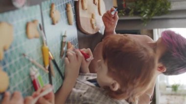 Küçük çocuklar ve annelerinin evde tatil hediyeleri hazırlarken renkli kremalarla Paskalya kurabiyelerini süsledikleri dikey çekim.