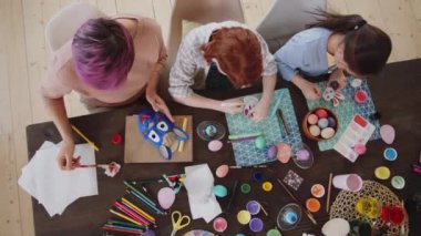 Küçük kızının, oğlunun ve annesinin resim ve el sanatları malzemeleriyle masada oturup dekoratif Paskalya oyuncakları yaparken en tepeden aşağı fotoğrafı.