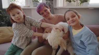 Mutlu anne, küçük kız ve oğul portresi evdeki kanepede sevimli bir köpek okşuyor, gülümsüyor ve kameraya poz veriyor.