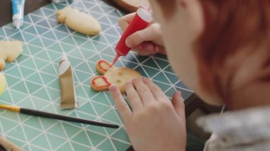 Kırmızı saçlı çocuğun el yapımı Paskalya kurabiyelerini kırmızı kremayla süslediği bir fotoğraf.