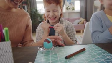 Paskalya yumurtalarını anne ve kız kardeşiyle birlikte oturma odasında süslerken gülen neşeli kızıl çocuk.