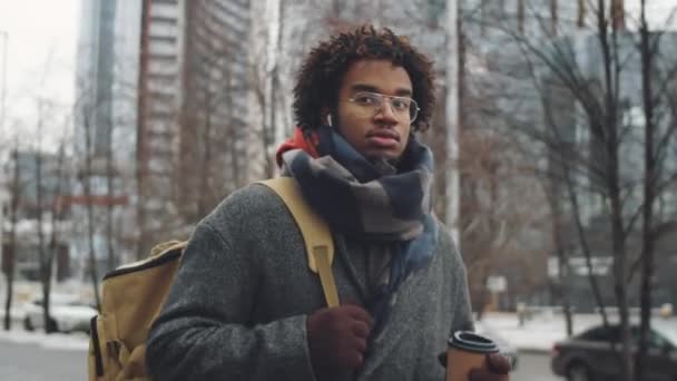 冬の日には 若いアフリカ系アメリカ人男性のメガネ ワイヤレスイヤフォン アウターのアークショットがコーヒーカップに行き 屋外で歩くために開催されます — ストック動画