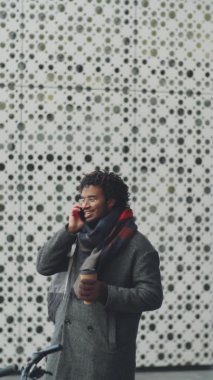 Tek kullanımlık kahve fincanı tutan ve cep telefonuyla konuşan genç bir Afro-Amerikalının dikey görüntüsü.