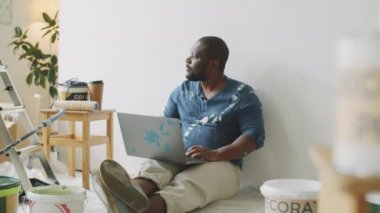 Giysileri boya lekeli orta boy Afro-Amerikalı bir adam dizüstü bilgisayarla yerde oturuyor ve evini yenilerken kameraya poz veriyor.