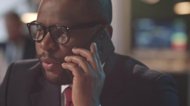 Resmi takım elbiseli ve gözlüklü Afro-Amerikan iş adamının ofiste çalışırken cep telefonuyla konuştuğu göğüs üstü fotoğraf.