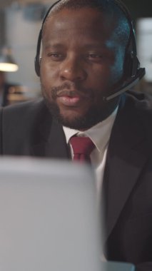 Kablosuz kulaklık ve resmi takım elbiseli Afro-Amerikan iş adamının ofisteki bilgisayardaki video konuşmasının dikey görüntüsü.