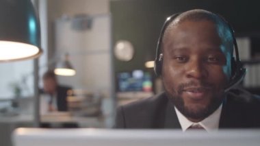 Kablosuz kulaklık ve forma giyen Afro-Amerikalı iş adamının ofis masasında oturup bilgisayardaki video konuşmasının görüntülerini izliyorum.