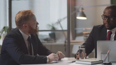 Resmi takım elbiseli Afro-Amerikalı iş adamı dizüstü bilgisayar kullanıyor ve ofis masasındaki beyaz erkek arkadaşla iş hakkında konuşuyor.