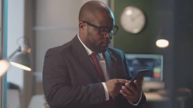 Resmi takım elbiseli siyahi işadamının belden yukarı vuruşu. Ofiste duruyor ve akıllı telefondan sosyal medyada kayıyor.
