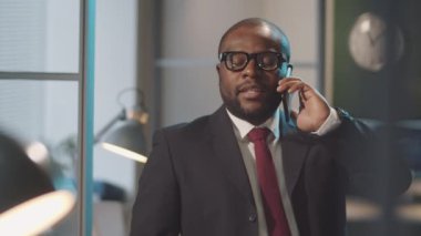Resmi takım elbiseli ve gözlüklü Afro-Amerikan iş adamının ofisinde cep telefonuyla konuşurken belini kaldır.