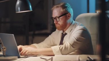 Resmi giyimli ve gözlüklü beyaz sakallı bir işadamı ofis masasında dizüstü bilgisayarda yazı yazıyor ve akşam mesai yaparken kameraya poz veriyor.