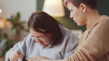 Down sendromlu yetişkin bir adam evde resim dersi sırasında kadın öğretmenle kağıt süsleme yaparken kağıt çiçeğe yapıştırıyor.