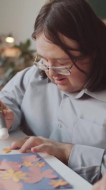 Down sendromlu bir adamın çiçek kağıdı yapıp kameraya gülümseyerek baktığı dikey çekim.