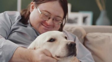 Down sendromlu neşeli bir adam gülümsüyor ve evdeki kanepede otururken Golden retriever köpeğini okşuyor.