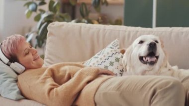 Kablosuz kulaklıklı neşeli kadın evdeki kanepede uzanıyor ve tatlı Golden Retriever köpeğini okşuyor.