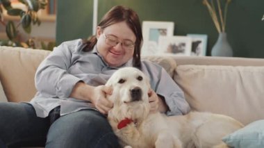 Down sendromlu mutlu adam evdeki kanepede oturuyor, gülümsüyor ve tatlı Golden Retriever köpeğini okşuyor.